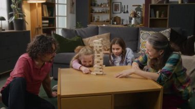 Orta boy sarışın beyaz bir kız tahta bloğu çekip kuleye yığılırken anne, engelli baba ve erkek kardeş izliyor ve tezahürat yapıyor.