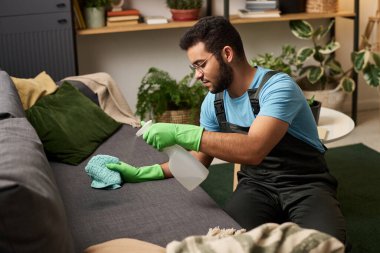Üniformalı bir adam ev işi sırasında odadaki kanepeyi temizlemek için deterjan kullanıyor.