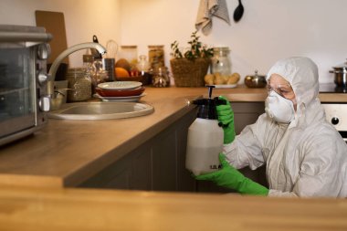 Koruyucu kıyafetli bir kadın mutfakta spreyle mobilyaları dezenfekte ediyor.
