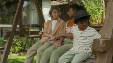 Orta yaşlı beyaz kadın ve melez Afrikalı Amerikalı çocuklar yaz günü yeşil bahçede salıncakta salıncakta gevşeyip, sohbet ediyor ve gülümsüyor.