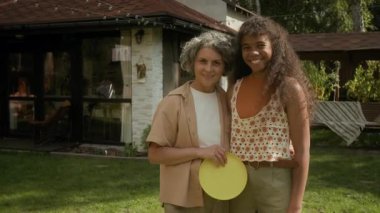 Mutlu, olgun, beyaz bir anne ve melez bir Afrikalı Amerikalı genç kızın orta boy portresi. Banliyö aile evinin yanındaki yeşil çimlerde yan yana duruyor, sarılıyor, kameraya bakıyor, gülümsüyor.