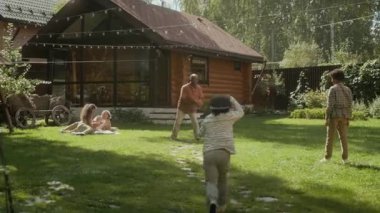 Siyah babanın genç oğlunu banliyö aile evinin yeşil çimlerinde beyzbol becerileriyle eğittiği, sonra da ona doğru koşan küçük çocuğu kucakladığı geniş açı. Aile sıcak yaz gününde birlikte dinleniyor.