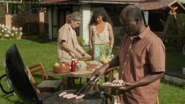 Orta boy Afro-Amerikan aile, bahçede barbekü partisine hazırlanıyor. Baba ızgaraya sosis koyuyor, anne-kız taze yemekle sofrayı kuruyor, oğullar akıllı telefondan oynuyor.