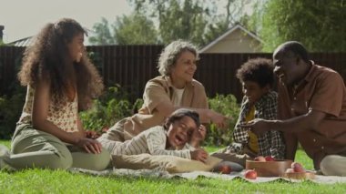 Beş Afrikalı Amerikalı baba, beyaz anne ve melez kızın bahçedeki çimlerin üzerinde uzanan, gülen, şakalaşan ve taze elma yiyen neşeli, çoklu etnik aileden oluşan yavaş çekim.