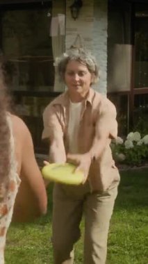 Orta boylu, orta yaşlı, beyaz bir kadının, aile evinin yeşil çimenliğinde kızıyla birlikte uçan diskle oynarken, hafta sonları birlikte dinlenirken çekilmiş dikey bir fotoğrafı.