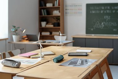 Eğitim malzemeleri ve arka plan kitaplıklarıyla dolu masaların olduğu bir sınıf. Diyagramlı yeşil karatahta akademik atmosfer ekler