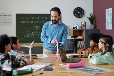 Öğretmen, bir grup özenli öğrenciye masada oturan yel değirmeni modelini anlatıyor. Eğitim araçları ve arka planda görsel yardımlarla dolu bir sınıf ortamı