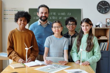 Sınıfta duran bir grup öğrenci ve öğretmen masanın üzerinde eğitim materyalleri ve arka planda bilimsel projelerle