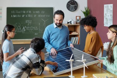 Sınıfta öğrencilere güneş panelleri ve rüzgar türbini modelleri kullanarak yenilenebilir enerji prensiplerini gösteren öğretmen. Eğitim posterleri ve saat, sınıf duvarlarını süslüyor