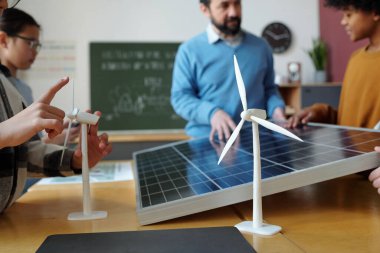 Sınıfta güneş paneli ve rüzgar türbini modelleri üzerinde çalışan bir grup öğrenci, arka planda öğretmenden rehberlik alarak yenilenebilir enerji projelerine odaklanıyorlar.