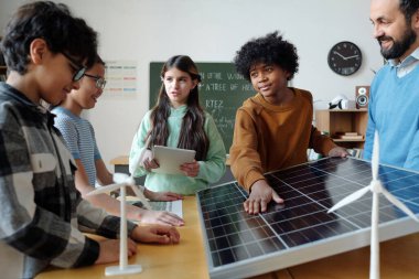 Bir grup öğrenci ve öğretmen sınıf ortamında yenilenebilir enerji projesi hakkında tartışıyorlar. Öğrenciler ve öğretmen çeşitli aygıtlar tutarken güneş paneli modelini inceliyor