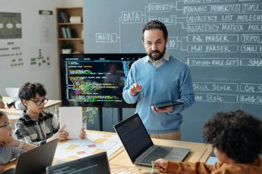Eğitimci elinde tablet tutarak çeşitli dizüstü bilgisayarlarla oturan öğrencilere programlama kavramlarını anlatıyor. Sınıfta kod ve diyagramlarla ilgileniyorlar.