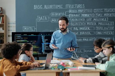 Öğretmen, masadaki dizüstü bilgisayarlarla ve ders kitaplarıyla oturan bir grup ilkokul öğrencisine kodlama kavramlarını anlatıyor. Sınıfta karatahta detaylı algoritmik yapılar yer alıyor.