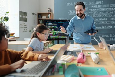Genç öğrencilerle birlikte sınıfa öncülük eden öğretmen derslerini aktif olarak takip ediyor. Sınıfta dizüstü bilgisayarlar ve çeşitli okul gereçleri kullanıyor.