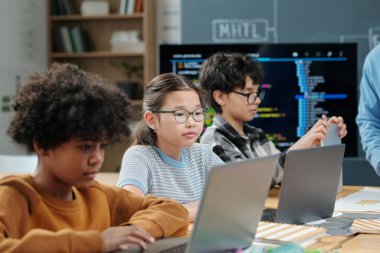 Sınıfta dizüstü bilgisayarlarla oturup çeşitli çizelgeler ve eğitim materyalleri sergileyen genç öğrenciler derslere odaklanıyorlar.