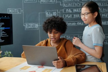 İşbirlikçi okul projesi üzerinde çalışan iki öğrenci, biri dizüstü bilgisayara odaklanmış, diğeri ise aktif olarak yardımcı oluyor. Arkaplanda diyagramları ve notları olan tebeşir tahtası