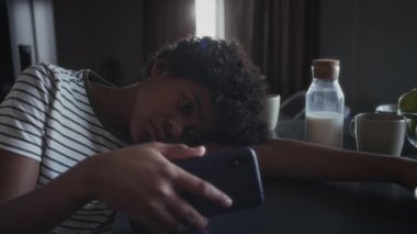 Orta ölçekte, kafası karışmış genç Afro-Amerikan çocuğun mutfak masasında yatıp, akıllı telefondan alaycı mesajlar okuması, zımbırtıyı bırakması, ağlaması ve gözlerini ovuşturması.