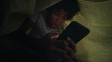 Orta karar, heyecanlı, 13 yaşında Afro-Amerikan bir çocuk gece yatakta, yorganın altında saklanıyor, akıllı telefondan uygunsuz ve eğlenceli içerikli görüntüler izliyor.