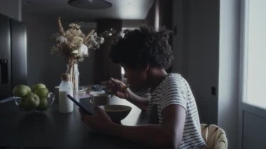 Genç Afrikalı Amerikalı bir çocuğun evde oturup kahvaltıda sütlü mısır gevreği yemesi sosyal medyaya bakması ve akıllı telefondan mesajları kontrol etmesi.