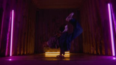 Sokak giysileri içinde, vücudunu döndürürken kucağına atlayan, neon ışığın altında modern break dans yapan yetenekli beyaz bir gencin zemin seviyesi yavaş çekimi.