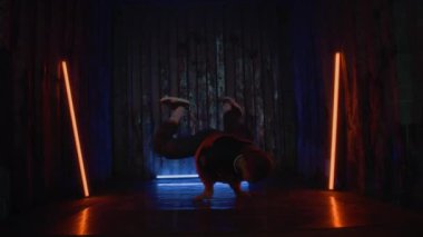 Yavaş çekimde, genç yetenekli beyaz çocuk amuda kalkarak hareketlerini sergiliyor, mavi ve turuncu neon ışıkların altında dans ediyor.