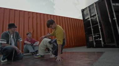 Tramvaylı, beyaz, erkek, yavaş çekim. Açık havada hip hop müzik dinliyor. Akranlarının önünde taş ve dondurucu hareketler yapıyor ve güneşli bir günde onu izliyor.