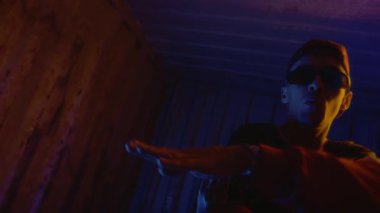 Gözlüklü, sokak tarzı kıyafetler ve gözlük takmış, gizemli neon ışıkların altında dans ederken kameraya bakan modaya uygun genç rapçinin el kameralı yavaş çekimi.