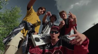 Şehir kıyafetleri içinde elinde retro kaset teybi olan dört beyaz gencin el kamerasıyla parlak güneşli bir günde açık havada hip hop el hareketleriyle kameraya baktıkları el kamerası görüntüsü.