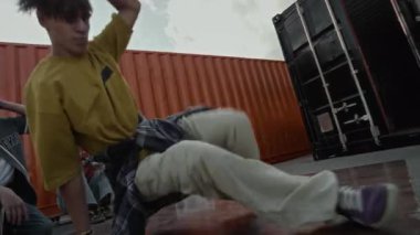 Sokak tarzı giyinmiş yetenekli beyaz çocuk dışarıda arkadaşlarının önünde nefes kesici break dans hareketleri sergiliyor ve yerde oturup onu izliyor.