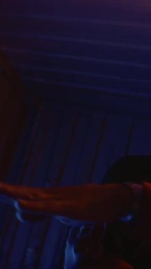 Gözlüklü, sokak tarzı kıyafetler ve gözlük takmış, gizemli neon ışıkları altında dans ederken kameraya bakan modaya uygun genç rapçinin dikey yavaş çekimi.