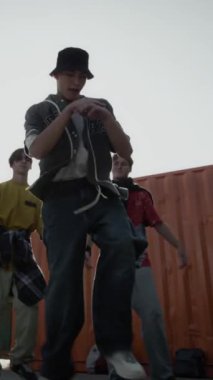 Kafkas jenerasyonu Z B-boy 'un arka planda akranlarının önünde break dans hareketleri yaparken, açık havada güneşli bir günde pratik yaparken çekilmiş dikey el çekimi.