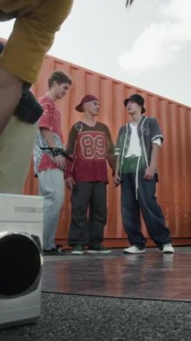 Sokak kıyafetleri içindeki beyaz erkek zoomerın dikey zemin görüntüsü retro kaset teybini açıyor ve açık hava güneşli bir günde akranlarının önünde breakdance yapıyor.