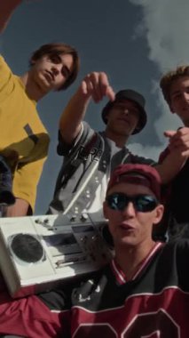 Güneşli bir günde açık havada hip hop el hareketleri yaparken ellerinde retro kaset teybi olan ve kameraya bakan dört beyaz gencin dikey düşük açılı portresi.