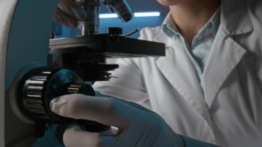 Laboratuvarda mikroskopla çalışan, beyaz önlüklü ve steril eldivenli kimliği belirsiz genç bilim adamının orta bölüm görüntüsü, arka planda erkek meslektaşı.