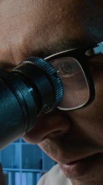 Laboratuvar ortamında mikroskop altında biyolojik örnekleri inceleyen gözlüklü genç erkek bilim adamının dikey yakın çekimi