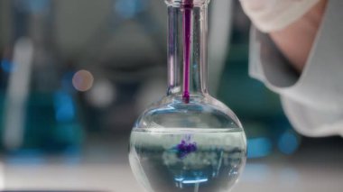 Bilinmeyen bir bilim adamının, mor maddeyi şeffaf sıvıyla şişeye doldurup deney sırasında sallamasının aşırı yakın çekimi.