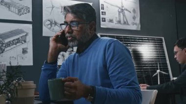 Tecrübeli Hintli erkek temiz enerji uzmanının cep telefonuyla görüşmesi sürdürülebilir teknolojiler hakkında danışmanlık yapması, modern mühendislik ofisinde kahve içmesi.