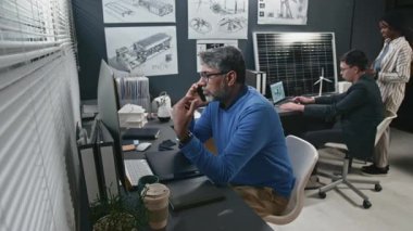 Üst düzey Hintli yenilikçi erkeğin bilgisayar monitörüne bakması ve modern ofisteki iş arkadaşlarıyla sürdürülebilir teknolojilerin gelişimini tartışması.