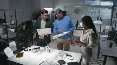 Modern ofiste multimedya ekranı ve güneş panelleri ile sürdürülebilir teknolojiler geliştirme konusunda fikir alışverişinde bulunmak için dizüstü ve teknik çizimler kullanan üç mühendisden oluşan tam çekim ekibi.