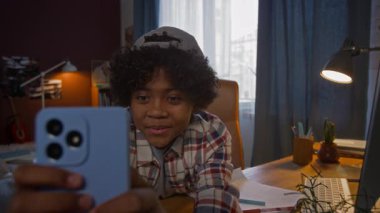 Sevimli Afro-Amerikan çocuk ödev sırasında masabaşında açık bir telif defteriyle otururken akıllı telefondan yeni video oyunu oynuyor.