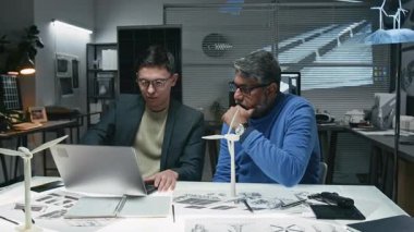 Genç ve olgun mühendislerin masada dizüstü bilgisayarlarıyla oturup ofiste yeni dijital rüzgar türbini modeli üzerinde çalışırken çekilmiş orta boy bir fotoğraf. Masada birden fazla teknik çizim ve iki yel değirmeni maketi var.