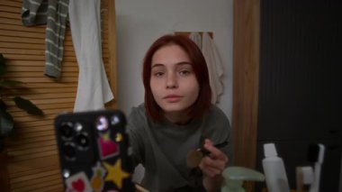 Güzel ergen kız yüzüne fırçayla pudra sürüyor, aynaya ve akıllı telefona bakıyor mesajlara cevap verirken ya da yeni şortları izlerken.