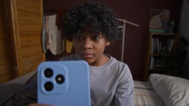 Sevimli Afro-Amerikan çocuk yatakta oturup akıllı telefon ekranına bakarken birden internetteki uygunsuz içeriği gördü, başını geriye çevirdi ve gülümsedi.