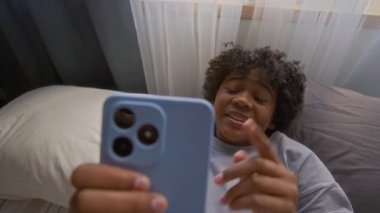 Sevimli, Afro-Amerikan tişörtlü, pencerede yatıp el kol hareketi yapan ve video sohbetinde arkadaşıyla iletişim kurarken akıllı telefon ekranına bakan tatlı bir öğrenci.