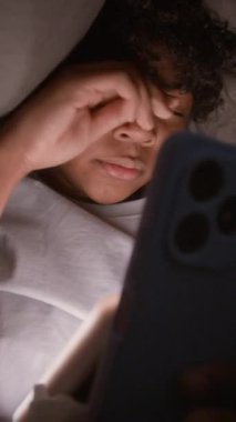 Uykulu öğrencinin karanlık bir odada yatarken gözlerini ovuşturup esnediği ve çevrimiçi video veya sosyal ağlardaki yeni paylaşımlarda gezindiği dikey bir fotoğraf.