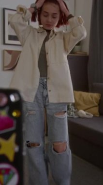 Gündelik giysiler içinde, akıllı telefon ekranında kayıt düğmesine basan ve oturma odasında kanepede dans etmeye başlarken çevrimiçi izleyiciler için yeni içerik oluşturan gençlerin dikey görüntüsü