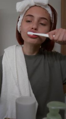 Dikey çekimde, beyaz yumuşak saç bandı takmış genç bir kız sabah banyoda aynanın önünde dişlerini fırçalarken ve akıllı telefondaki yeni online şortlara bakarken.