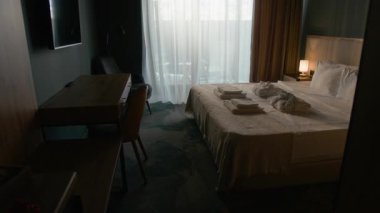 Temiz havlular ve katlanmış bornozlarla dolu rahat otel odasının içinde düzgün yapılmış yatak, sandalye ve geniş pencereli masa.