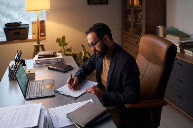 Formalite ve gözlük takan sakallı bir adam ofisinde oturmuş hastalar hakkında bilgi topluyor.