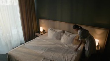 Otel hizmetçisinin yatak odasına beyaz çarşaf ve yastıklar yerleştirdiği, başucu lambaları ve battaniyenin üzerine katlanmış havlular yerleştirdiği yüksek açılı bir manzara.